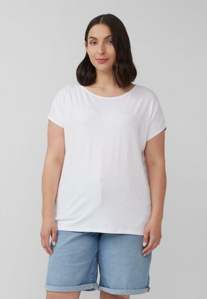 Vrouw met schouderlang donker haar, gekleed in een eenvoudige witte t-shirt en lichtblauwe denim shorts, tegen een grijze achtergrond.