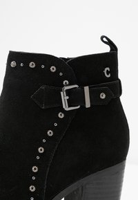 Botín de ante negro con tachuelas plateadas decorativas y una hebilla. El botín tiene un tacón grueso y un lazo de tela en la parte trasera.