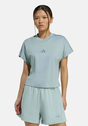 Jonge vrouw die staat, gekleed in een lichtblauw Adidas topje met korte mouwen en bijpassende korte broek, met zichtbare Adidas-logo's op beide.