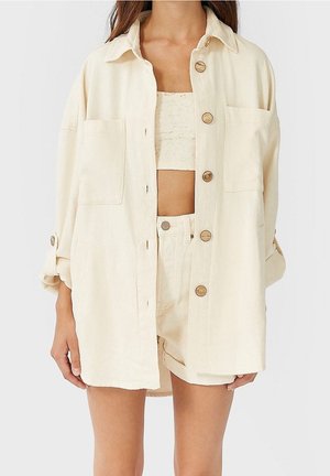 Femme portant une chemise beige oversize boutonnée, un short taille haute assorti et un crop top en dentelle, debout les mains détendues le long du corps.