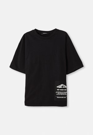 T-shirt noir surdimensionné en coton, avec des manches courtes, un col rond et une étiquette avec du texte blanc sur une poche latérale.