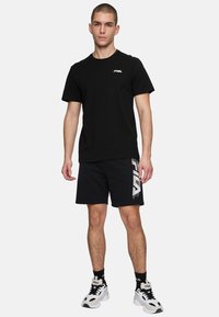 Musta lyhythihainen t-paita, jossa on pieni logo, yhdistettynä mustiin shortseihin, joissa on valkoinen brändigrafiikka, ja valkoisiin tennareihin mustilla yksityiskohdilla.