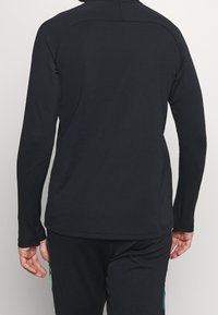 Musta pitkähihainen paita korkealla kaula-aukolla, sileää kangasta, litteät saumat ja istuva muotoilu. Minimalistinen tyyli ilman näkyviä logoja.