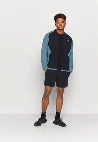 Black athletic jacket with teal accents, zip front, paired with black shorts. Features lightweight fabric and fitted design. Black sneakers.