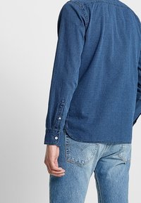 Muž v modré texturované košili s dlouhým rukávem a světle modrých džínách, zobrazený zezadu s rukou uvolněnou po boku.