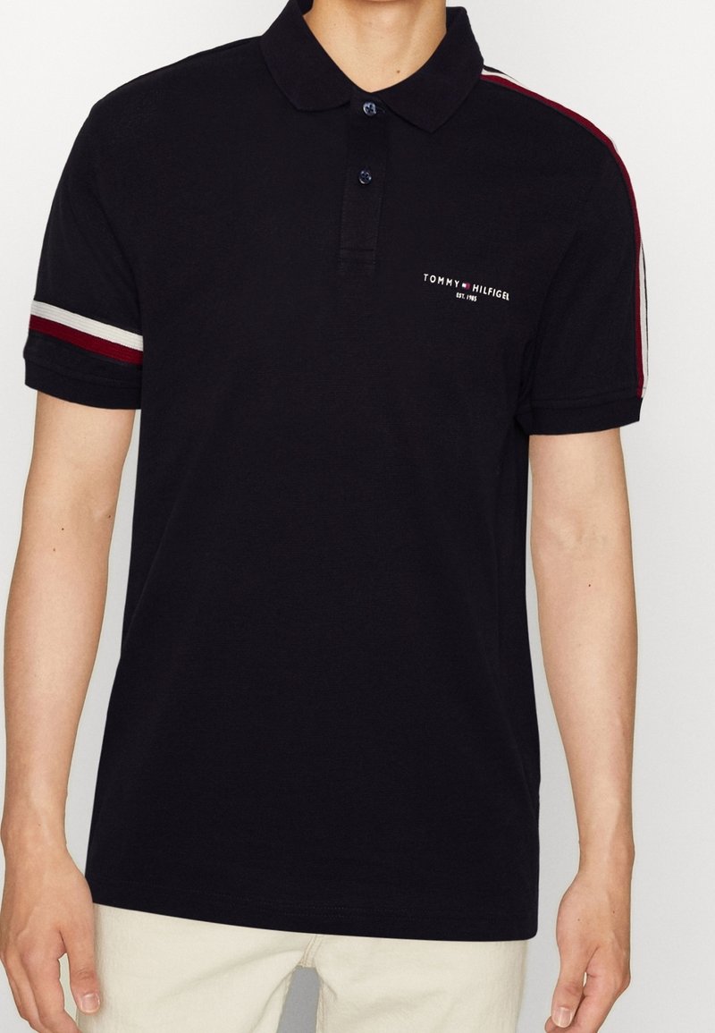 Homme portant un polo noir à manches courtes avec des rayures rouges et blanches sur les manches et le logo Tommy Hilfiger sur la poitrine.