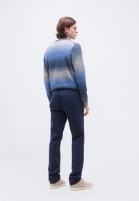 Νεαρός άνδρας όρθιος, φοράει πουλόβερ σε αποχρώσεις του μπλε, σκούρο μπλε παντελόνι και μπεζ αθλητικά παπούτσια, κοιτάζοντας προς τα πλάγια με φόντο ένα απλό σκηνικό.