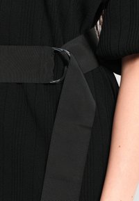 Robe en tissu côtelé noir avec une large ceinture en tissu noir fixée à la taille d'une personne par une boucle double en métal en forme de D.