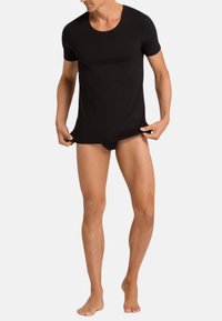 Man wearing a fitted black short-sleeve shirt and black briefs, holding the bottom hem of the shirt with both hands.