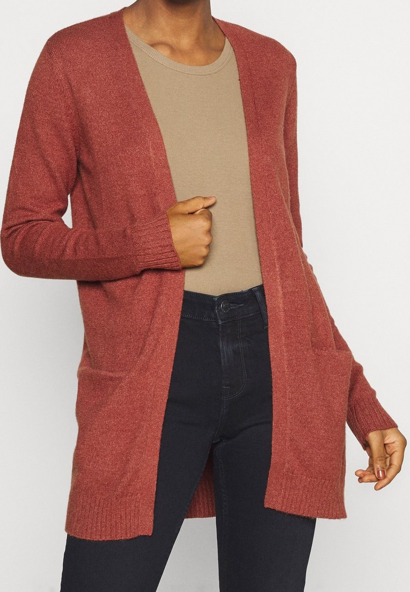 Personne portant un cardigan ouvert couleur rouille par-dessus une chemise beige et un pantalon noir taille haute, main tenant le bord du cardigan.