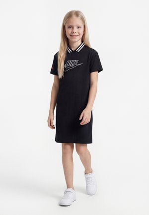 Fiatal lány áll, fekete Nike ruhát visel, fehér csíkozású gallérral és fehér tornacipővel, egyszínű fehér háttér előtt.