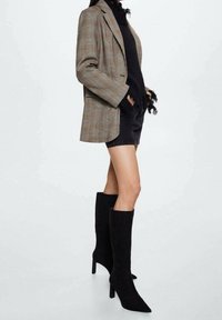 Bottes noires en daim montantes avec un bout pointu et un talon aiguille, associées à un blazer à carreaux et un top noir avec des détails en franges.