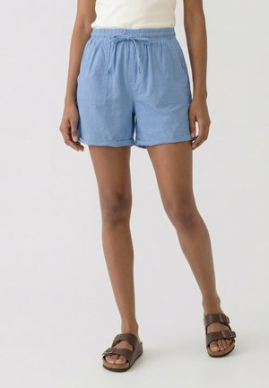 Person, die hellblaue Shorts mit Kordelzug, ein weißes Oberteil und braune Sandalen mit Schnalle trägt, steht vor einem einfarbigen hellen Hintergrund.