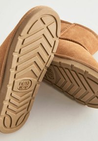 Tan mockasuinsboots med en texturerad gummisula som har ett fiskbensmönster och en präglad logotyp. Synlig sömnad längs ovandelen.