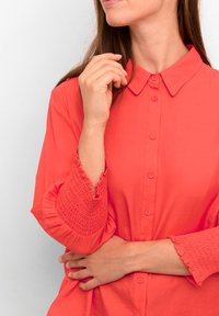 Donna che indossa una vivace camicia corallo con bottoni e polsini arricciati, con un braccio incrociato e l'altra mano vicino al collo.