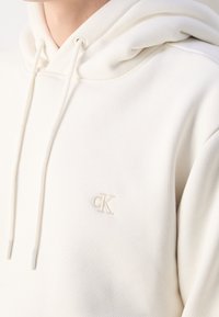 Gros plan d'une personne portant un sweat à capuche crème avec un cordon de serrage et un petit logo "cK" brodé sur la poitrine.