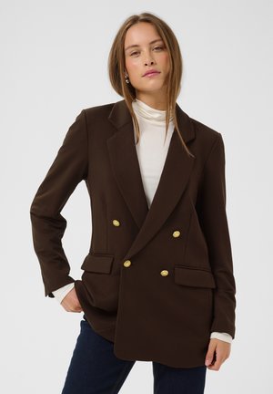 Junge Frau trägt dunkelbraunen, doppelreihigen Blazer mit goldenen Knöpfen, weißen Rollkragenpullover und dunkle Jeans, steht vor hellem, einfarbigem Hintergrund.