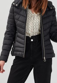 Veste en duvet noire avec capuche, présentant une texture matelassée, une fermeture éclair argentée et un accent en métal rond. Portée sur un blouse à motifs et un pantalon noir.