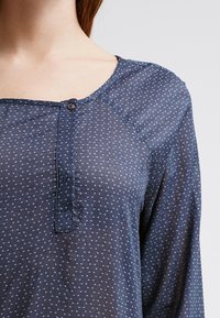 Námořnická modrá blůza s geometrickým vzorem, na kterém jsou malé světle modré trojúhelníky. Tkanina vypadá jemně, má kulatý výstřih a detaily s knoflíky.