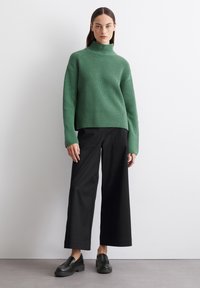 Grüner gerippter Rollkragenpullover kombiniert mit schwarzen weiten Hosen und schwarzen Slippers. Der Pullover hat eine lässige Passform und lange Ärmel.
