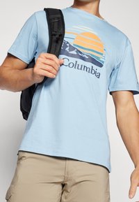 Hombre que lleva una camiseta ligera azul de Columbia con un gráfico de montaña y sol, pantalones de color beige y una correa de mochila negra sobre el hombro.