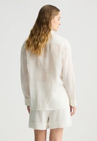 Camicia bianca in lino con maniche lunghe e orlo curvo, abbinata a pantaloni corti bianchi coordinati. Il tessuto sembra leggero e leggermente strutturato.