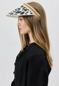 Junge Frau in schwarzem, knopfgebendem Hemd, die eine weiß-schwarze Tierdruck-Visor mit beigen Streifen trägt und nach links in einem Studio-Setting blickt.