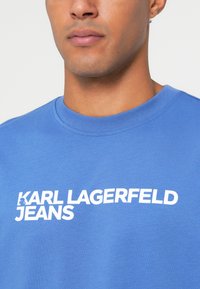 Sininen puuvillaneule, jossa on pyöreä kaula-aukko ja valkoiset tekstit "KARL LAGERFELD JEANS" rinnassa rohkealla, modernilla fontilla.