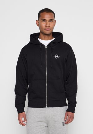 Junger Mann trägt einen schwarzen Zip-Up-Hoodie mit Replay-Logo und hellgraue Jogginghose, steht vor einem schlichten Hintergrund.