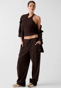 Hnědý krátký top bez rukávů, volné hnědé kalhoty se šňůrkou a odpovídající oversized košile. Hladká textura s minimálními doplňky.