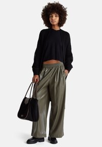 Kobieta o kręconych włosach, ubrana w czarny sweter, oliwkowe szerokie spodnie oraz czarne buty, trzymająca dużą czarną torebkę na białym tle.
