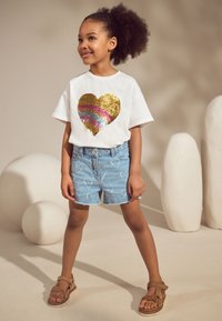 Fehér póló arany és szivárvány színű flitteres szív mintával, világoskék farmer rövidnadrággal, mely fehér szív mintákat visel, és barna szandállal.