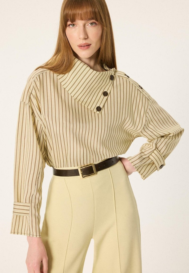 Femme aux longs cheveux roux portant un chemisier beige rayé boutonné et un pantalon taille haute jaune clair avec une ceinture noire, posant avec la main sur la hanche.