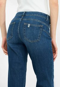 Jeans in denim blu con gamba dritta, dotati di due tasche posteriori e un'etichetta metallica. Il tessuto ha una consistenza liscia e una leggera scoloritura.