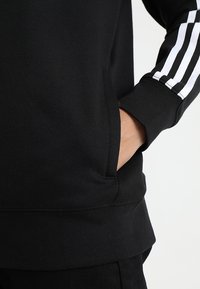 Svart sweatshirt med vita randiga detaljer på ärmarna, ribbad nederkant och ärmslut samt en sidoficka. Mjuk, strukturerad tyg.