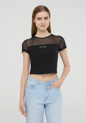 T-shirt nera cropped con maniche in rete trasparente, caratterizzata da un testo ricamato "falo' per la trama" sul petto. Abbinata a jeans azzurri chiari.