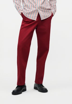 Homme portant un pantalon slim rouge foncé, des chaussures habillées noires et une chemise blanche rentréee avec des rayures verticales rouges et bleues.