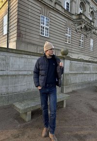 Mees, kes kannab beeži mütsi, musta paksud jope, tumedaid teksaseid ja pruune saapaid, seisab kiviaia ja mitme aknaga kaunilt ehitud hoone lähedal.