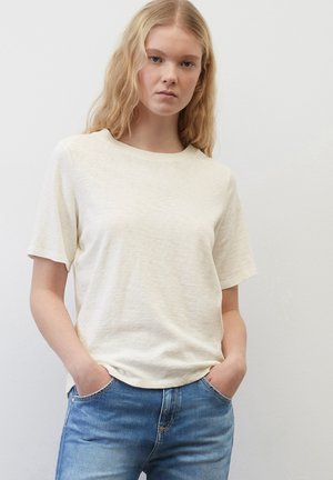 Tricou scurt cu mânecă scurtă, de culoare bej deschis, dintr-un material moale și texturat, cu un decolteu rotund. Purtați cu blugi albaștri și mâinile în buzunare.