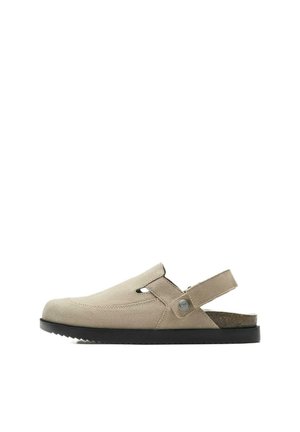 Béžová semišová slip-on sandála s čiernou podrážkou, nastaviteľným zadným remienkom so zapínaním na gombík a uzavretou špičkou na bielom pozadí.