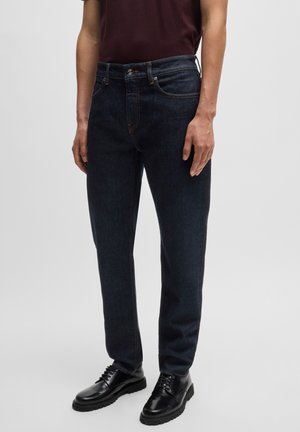 Dark blue denim jeans with a straight-leg design, contrasting orange stitching, and a classic five-pocket style, paired with black shoes.