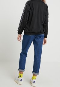 Svart idrottsjacka med vita randiga detaljer, blå denim jeans uppvikta samt vita sneakers. Gula strumpor är synliga ovanför skorna.