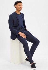 Granatowy, dopasowany garnitur z taliowanym żakietem i wąskimi spodniami, zestawiony z granatową koszulą i ciemnymi butami wsuwanymi. Gładka struktura.