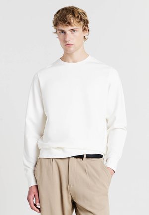 Weißer Baumwollpullover mit Rundhalsausschnitt und langen Ärmeln, mit lockerem Schnitt und gerippten Bündchen. Kombiniert mit beigen Hosen.