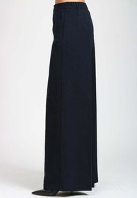 Donna che indossa pantaloni a vita alta e a gamba larga di colore blu navy, abbinati a scarpe con tacco a punta nere, mostrata di lato su uno sfondo bianco semplice.