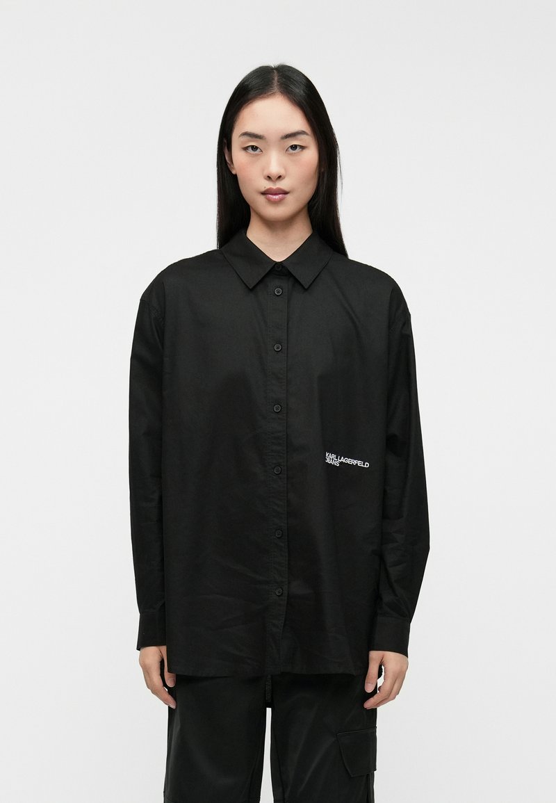 Camisa negra de manga larga con botón en la parte delantera, que cuenta con un cuello puntiagudo y un logo bordado en el pecho izquierdo. Textura de tela suave.