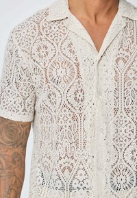 Chemise à manches courtes en dentelle couleur beige clair avec un motif ajouré complexe, dotée d'un col classique et d'une fermeture à boutons.
