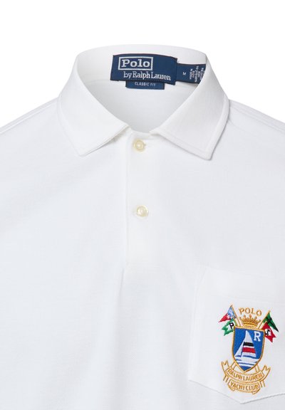 Polo classique blanc Polo Ralph Lauren à coupe classique avec deux boutons et un écusson brodé multicolore du Yacht Club sur la poche poitrine.
