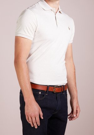 Homme portant un polo blanc à manches courtes, un jean bleu foncé et une ceinture en cuir marron avec une boucle en laiton.