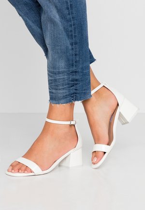 Białe sandały na obcasie typu blok z kwadratowym kształtem, paskiem wokół kostki oraz dwoma szerokimi paskami na palce. Gładkie wykończenie i minimalistyczny design.